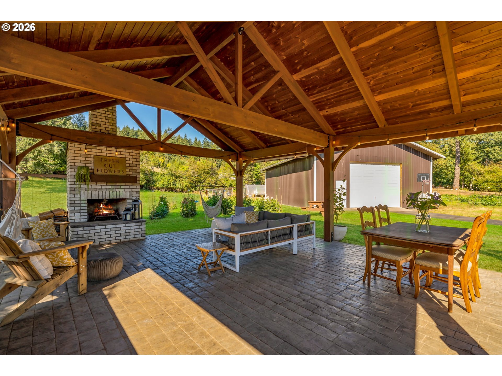 Covered patio with timber beams, brick fireplace, lounge seating, and dining table beside a detached garage.