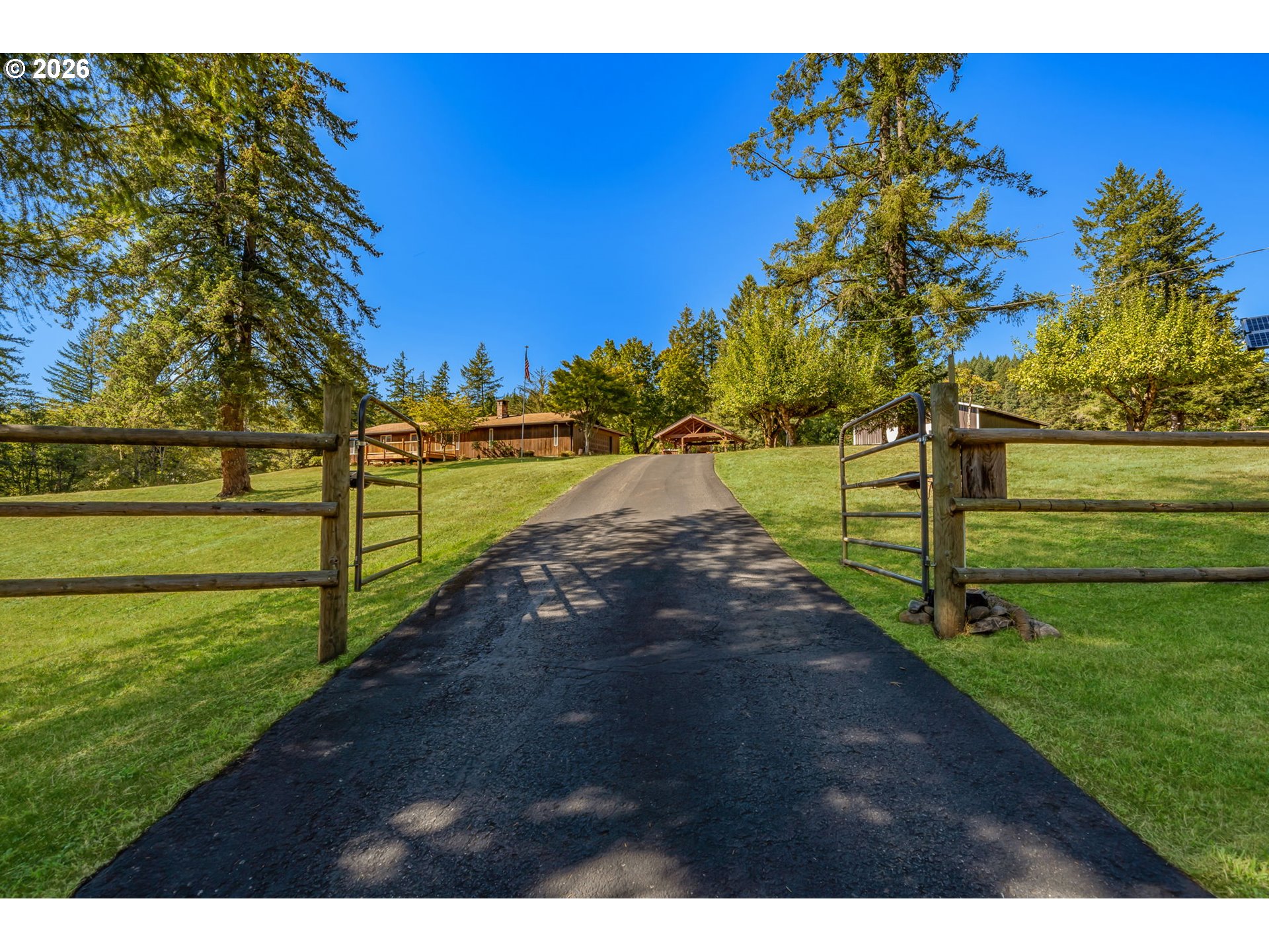 Paved driveway through open wooden gates leads uphill across a lawn to buildings among tall trees.