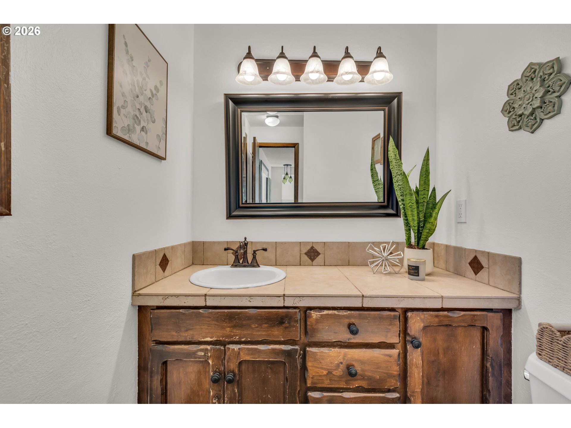 Bathroom vanity with a tiled countertop, rustic wood cabinets, and a large framed mirror.