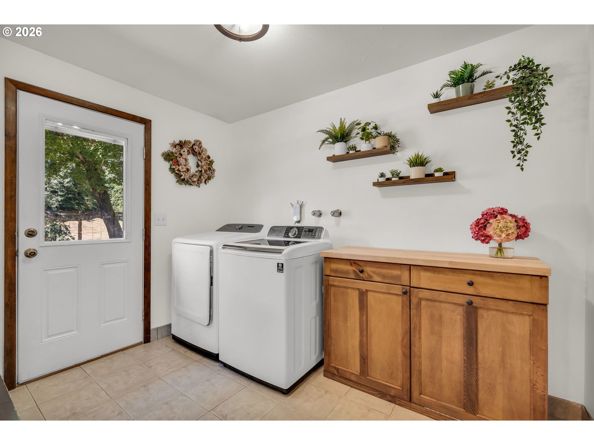Laundry room with white washer and dryer, wooden storage cabinet, and plant-lined floating shelves.