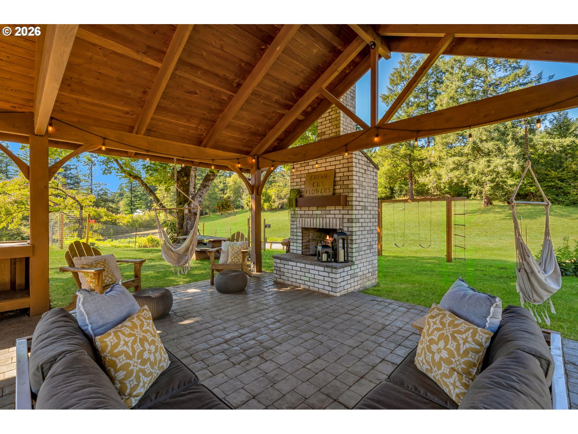 Covered patio with a brick fireplace, cushioned seating, and hanging chairs overlooking a green lawn.