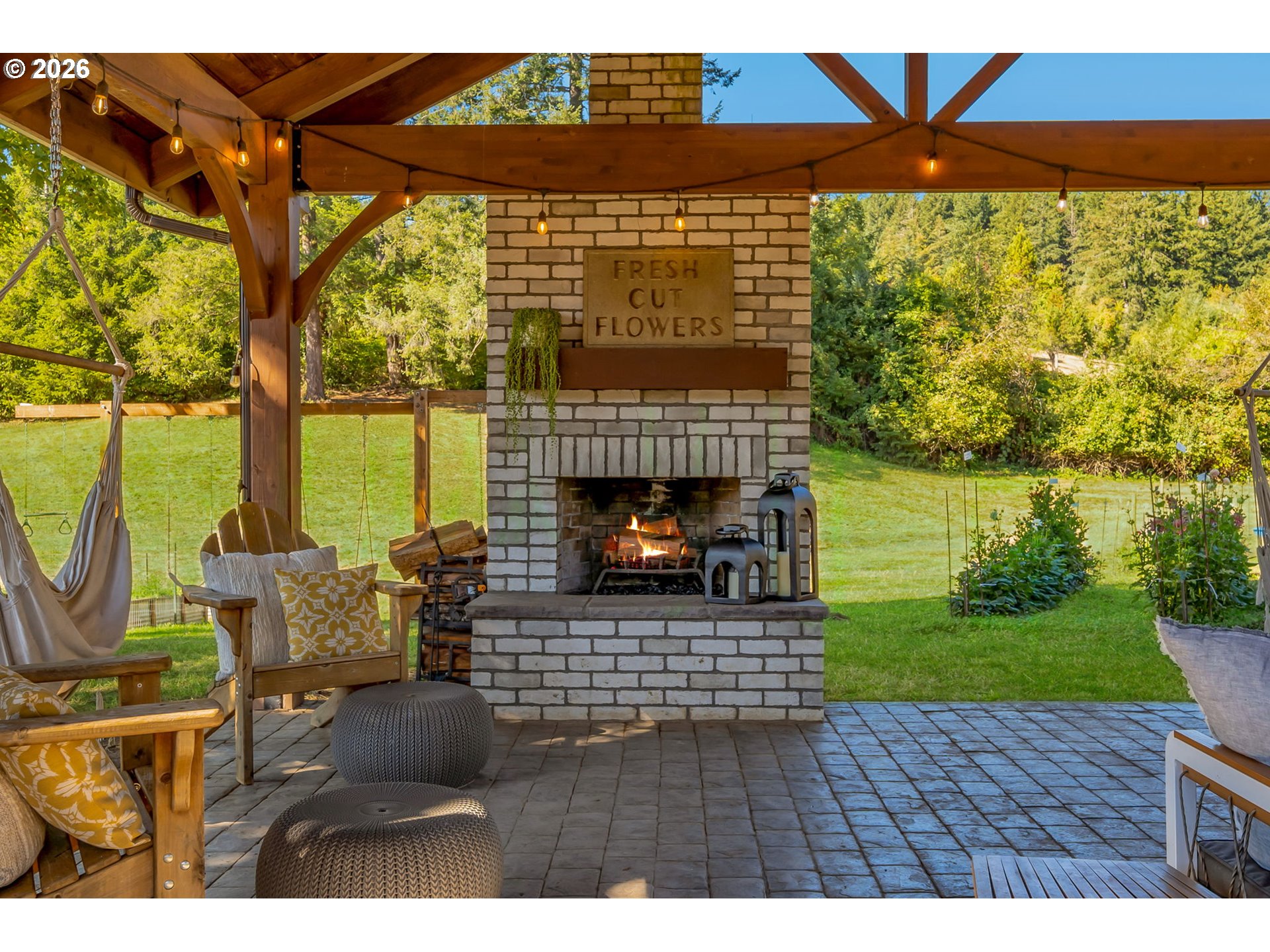 Covered patio with a brick fireplace, timber roof, and seating overlooking a wooded lawn.