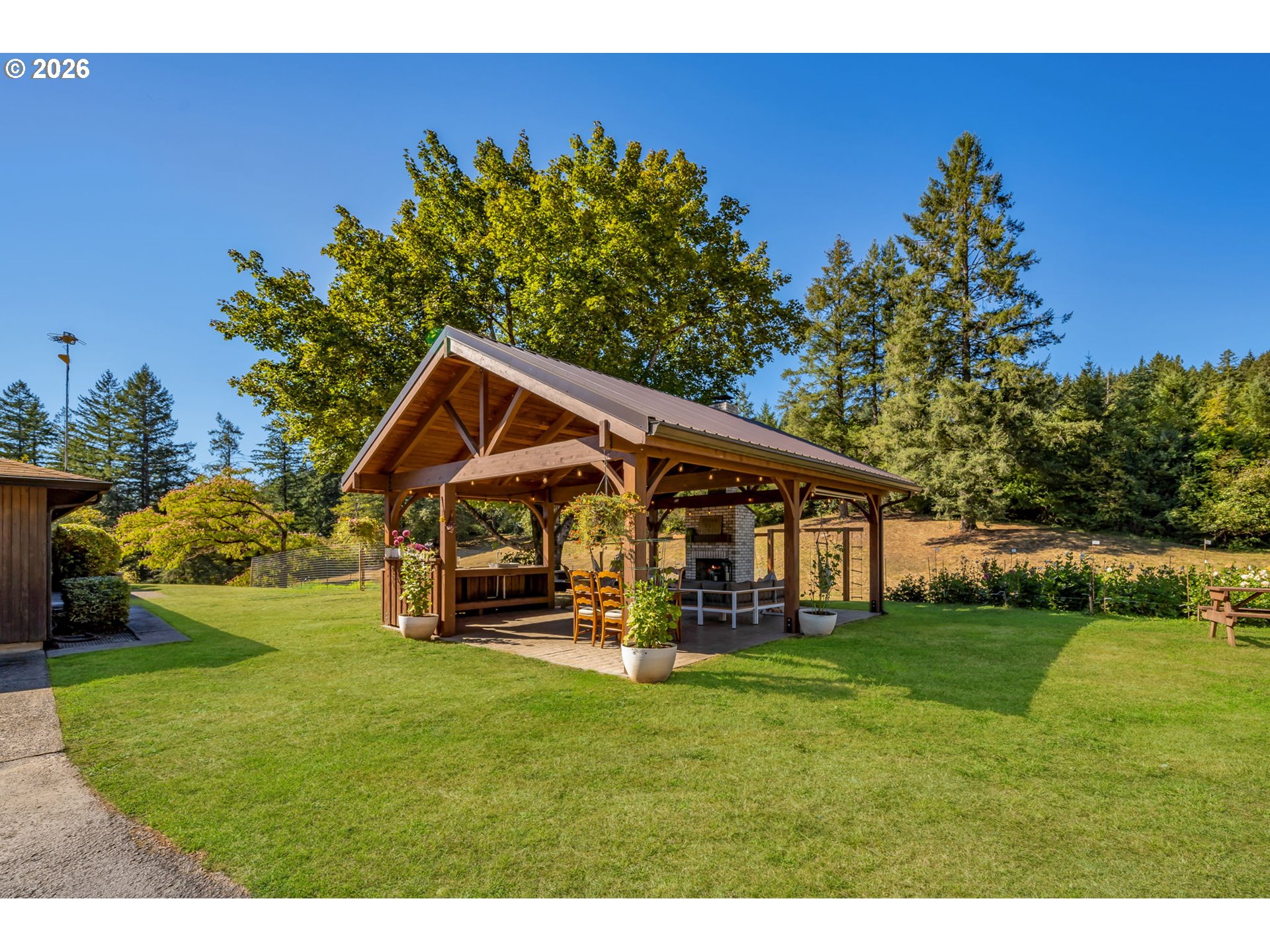 Covered timber pavilion with a brick fireplace and dining furniture on a landscaped lawn.
