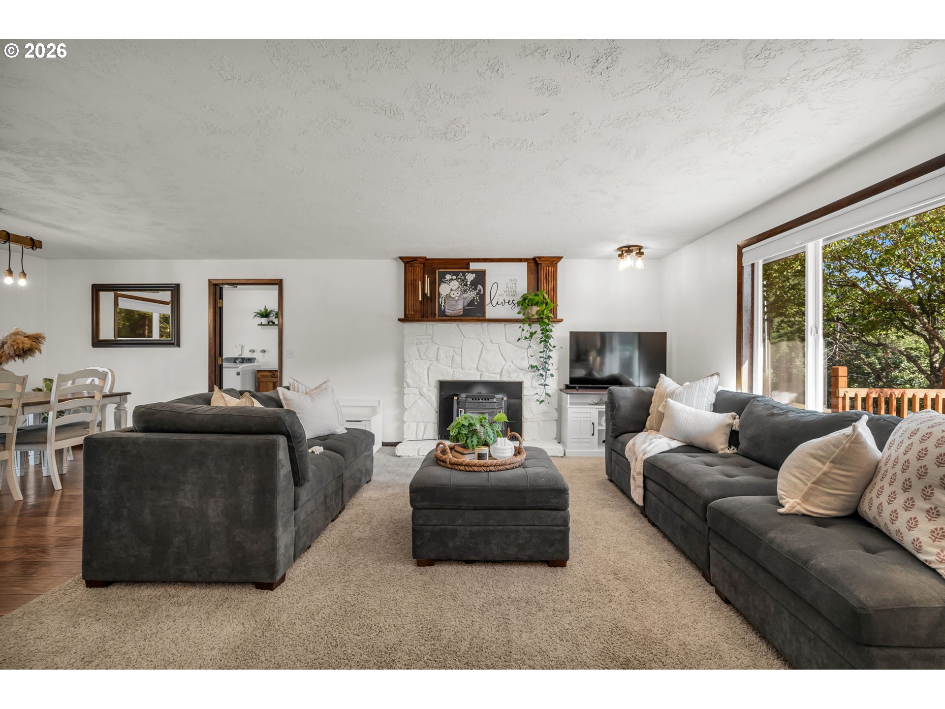 Living room with dark sofas, a white stone fireplace, and a large window overlooking leafy trees.