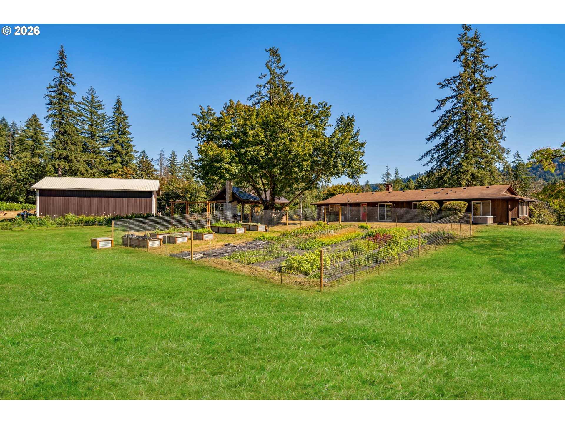Single-story home and outbuilding beyond a fenced vegetable garden and broad green lawn.