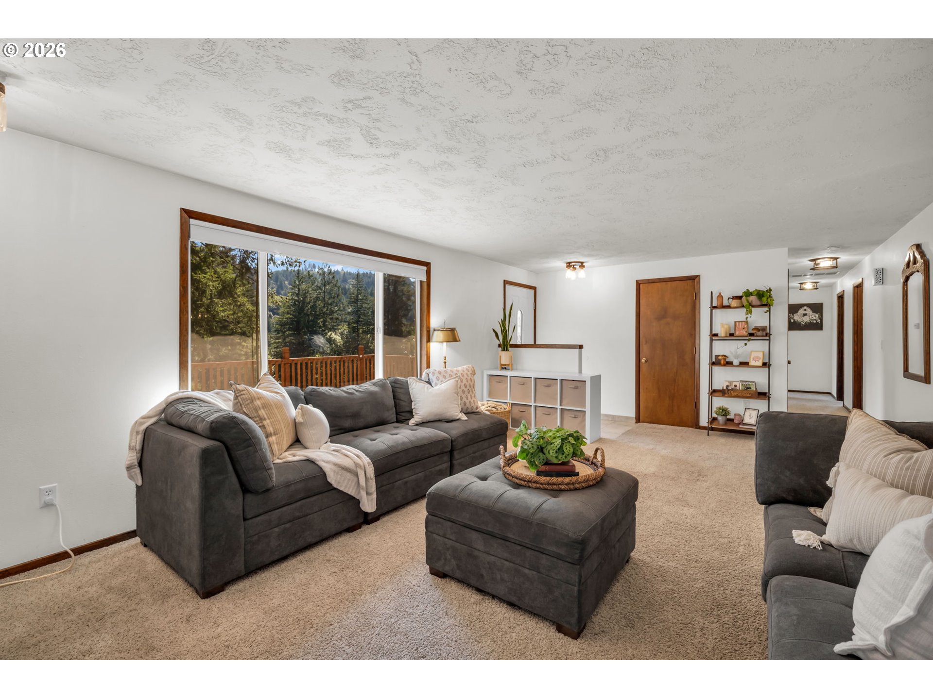 Living room with gray sofas and ottoman beside a large window overlooking a wooded hillside.