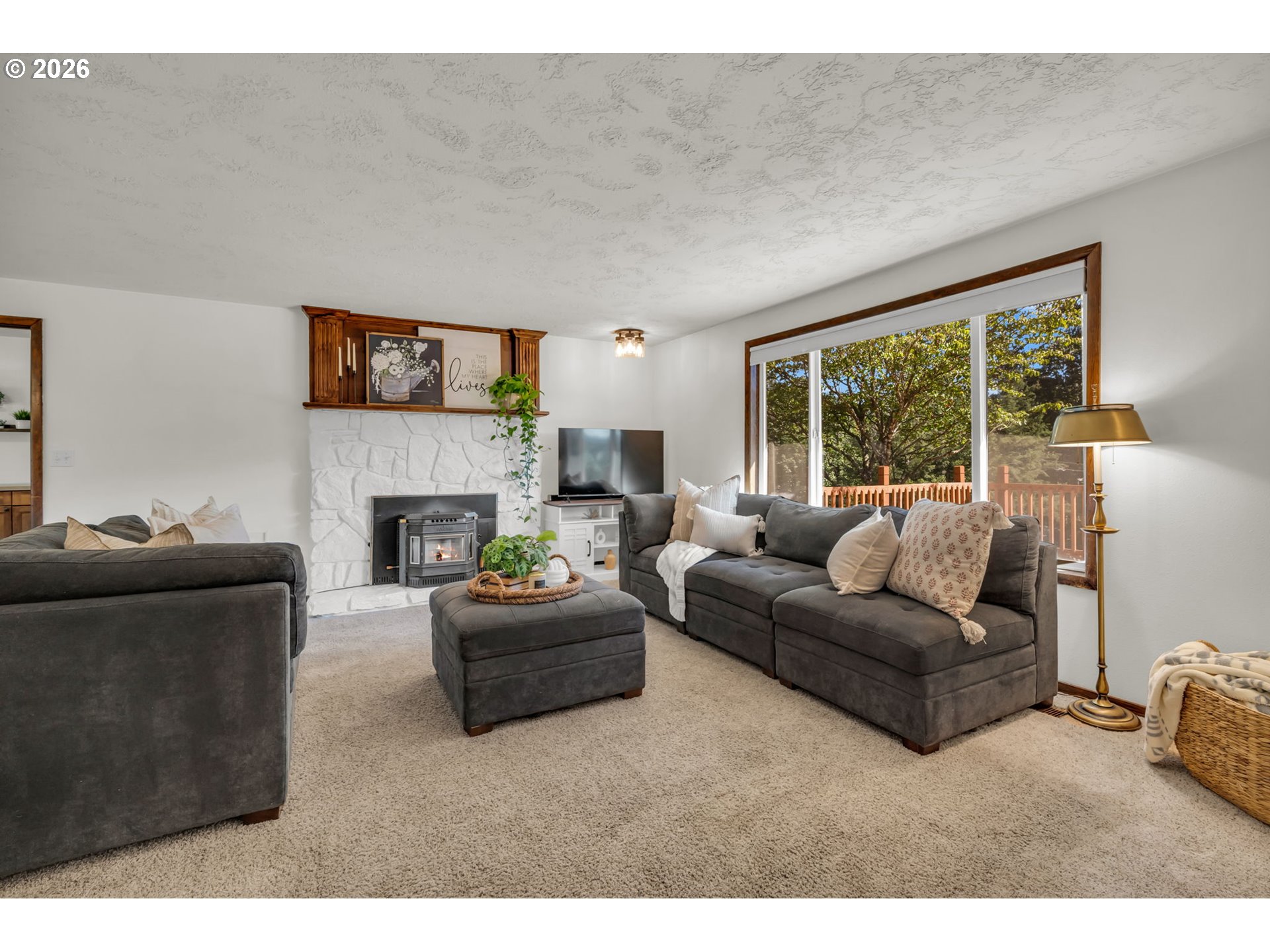 Living room with gray sectional sofas, a stone fireplace and wood stove, and a large window overlooking trees.