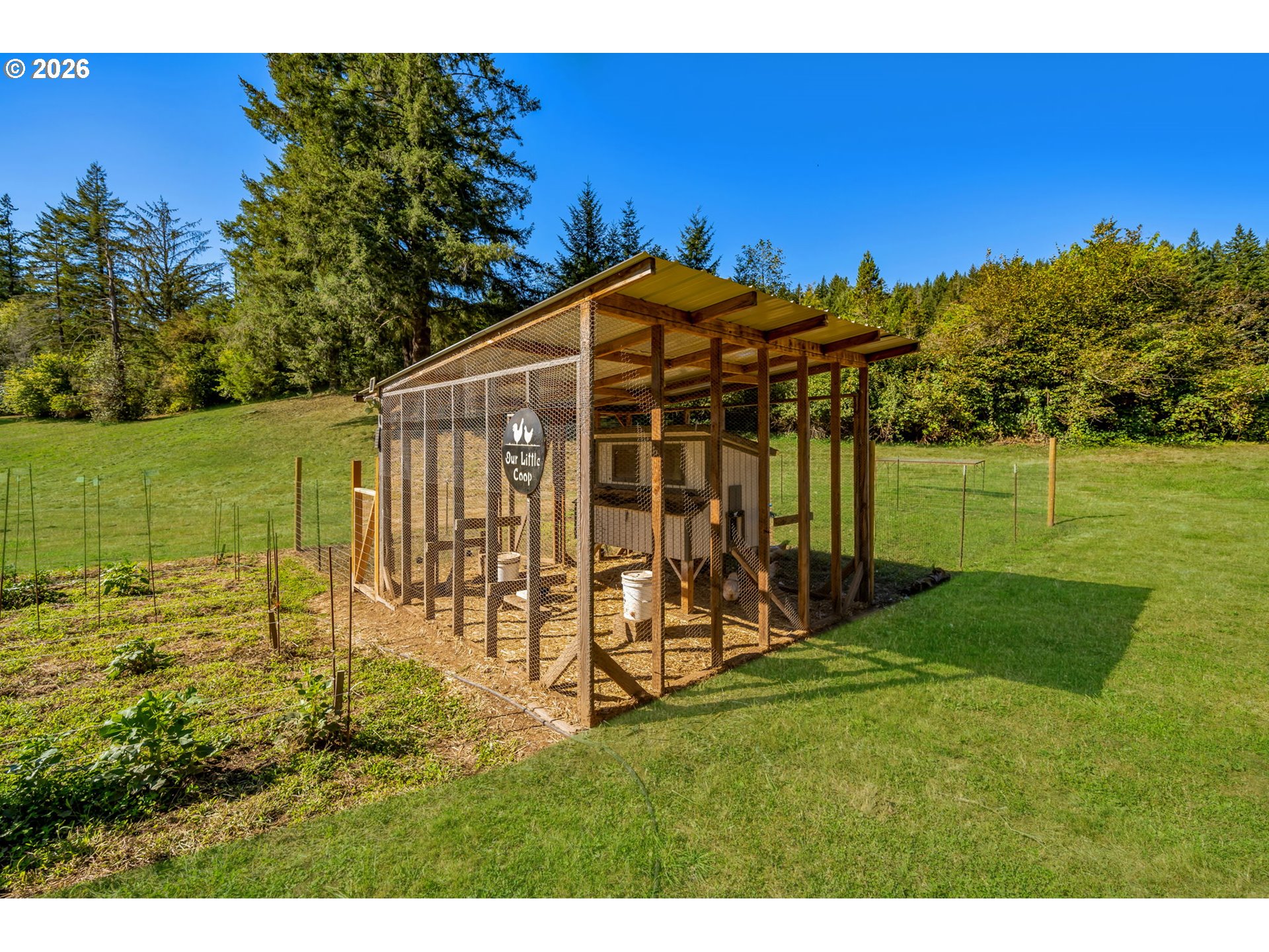 Wire-mesh chicken coop with a covered wooden roof and a sign reading “Our Little Coop.”