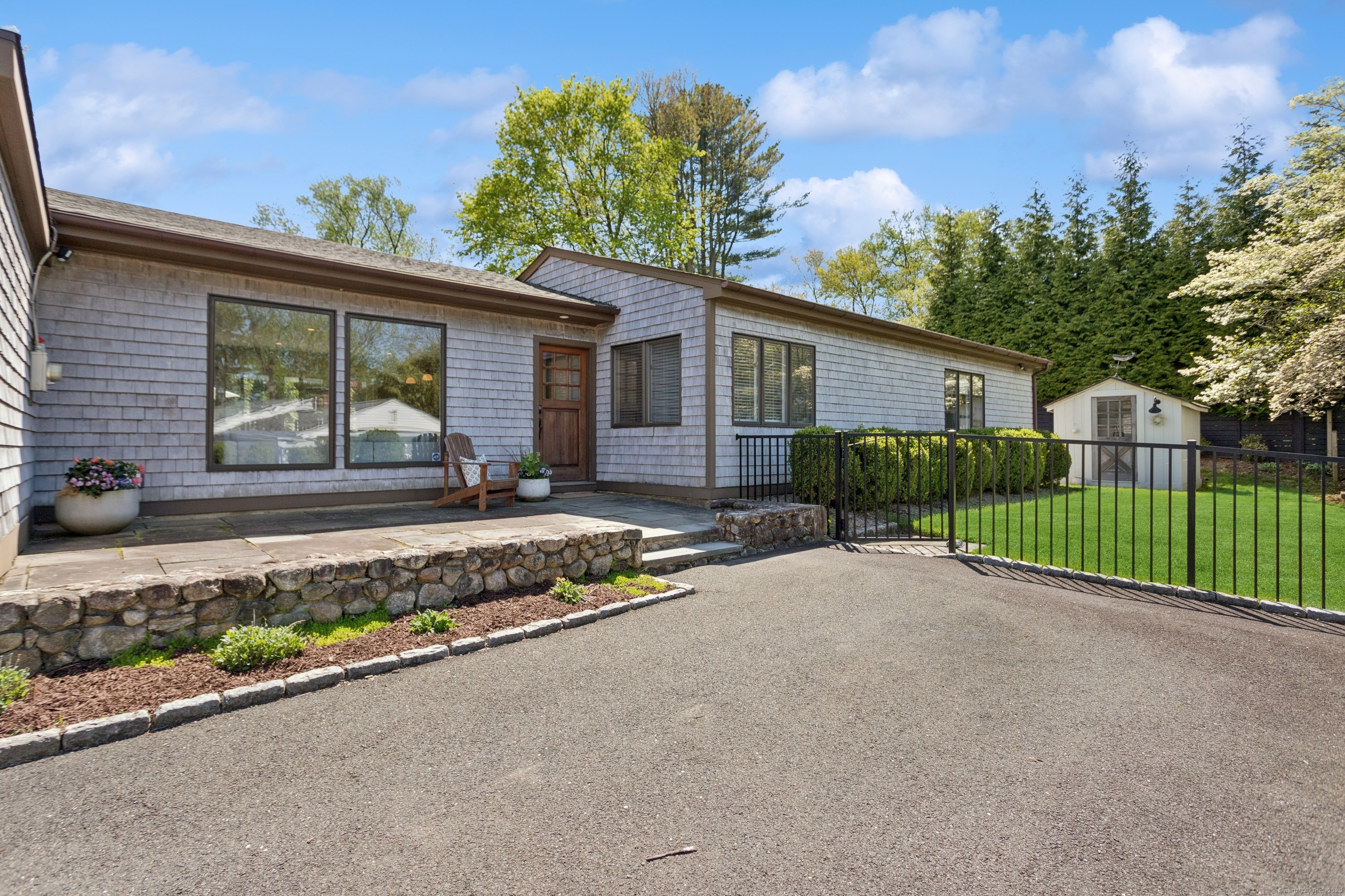 Gray-shingle home with a stone-edged patio, black metal fence, green lawn, and detached shed.