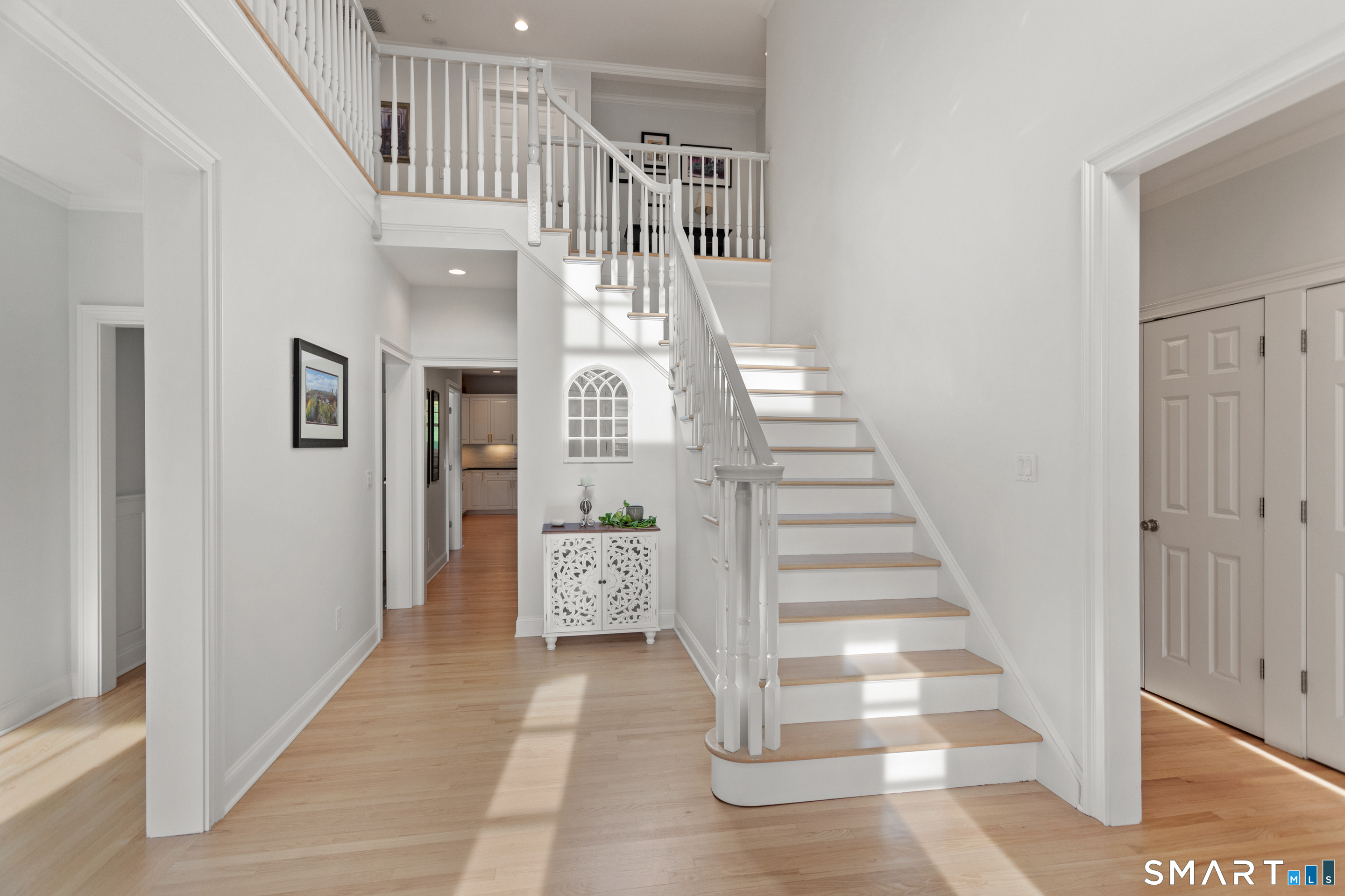 Bright foyer with a white staircase, light wood treads, hardwood floors, and a decorative cabinet beneath an arched window.