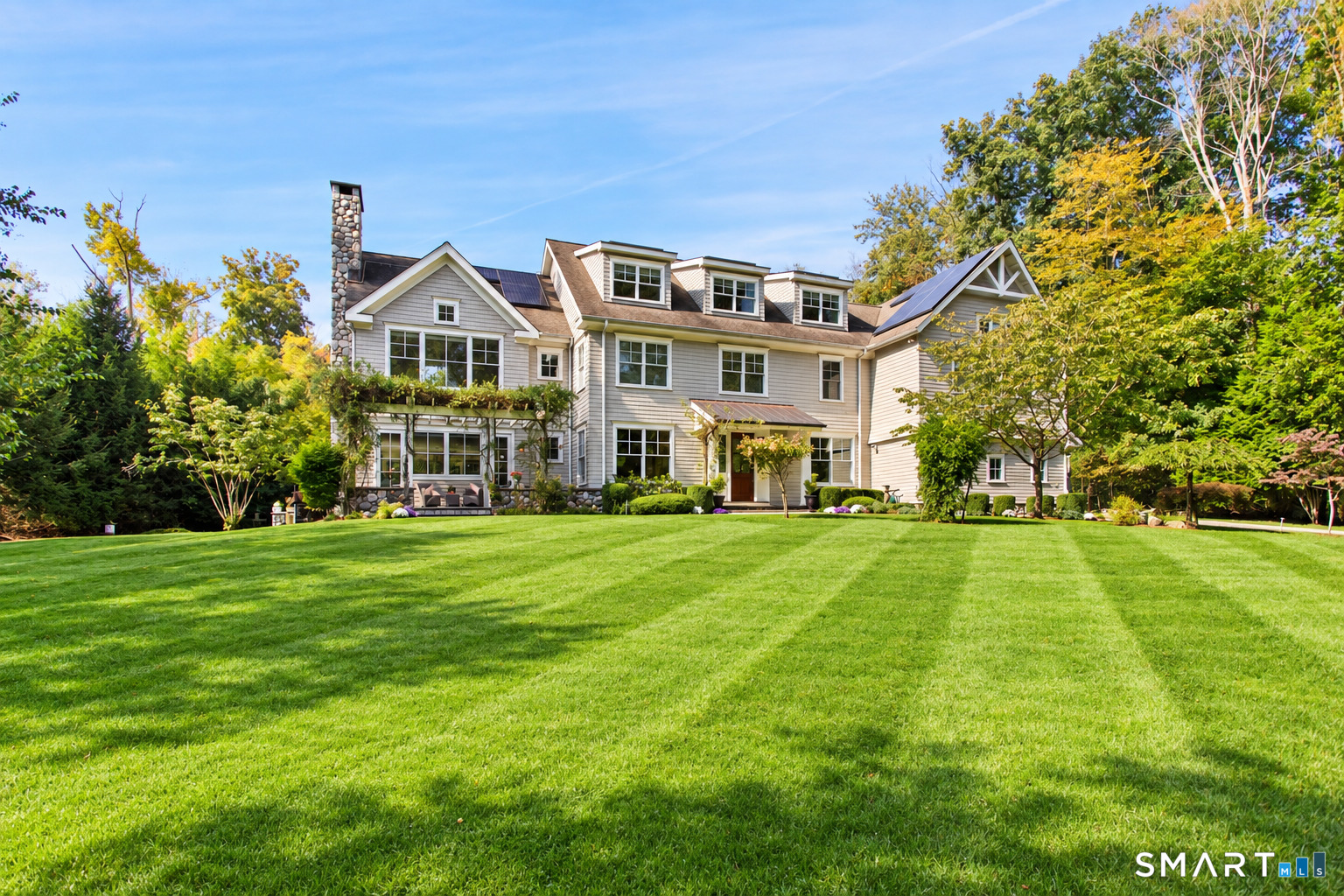 Large light-colored house with solar panels and stone chimney behind a broad green lawn.