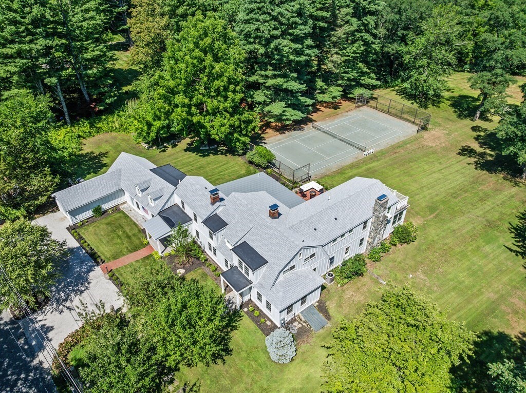 Aerial view of a large gray-shingled home with a fenced tennis court and wooded grounds.