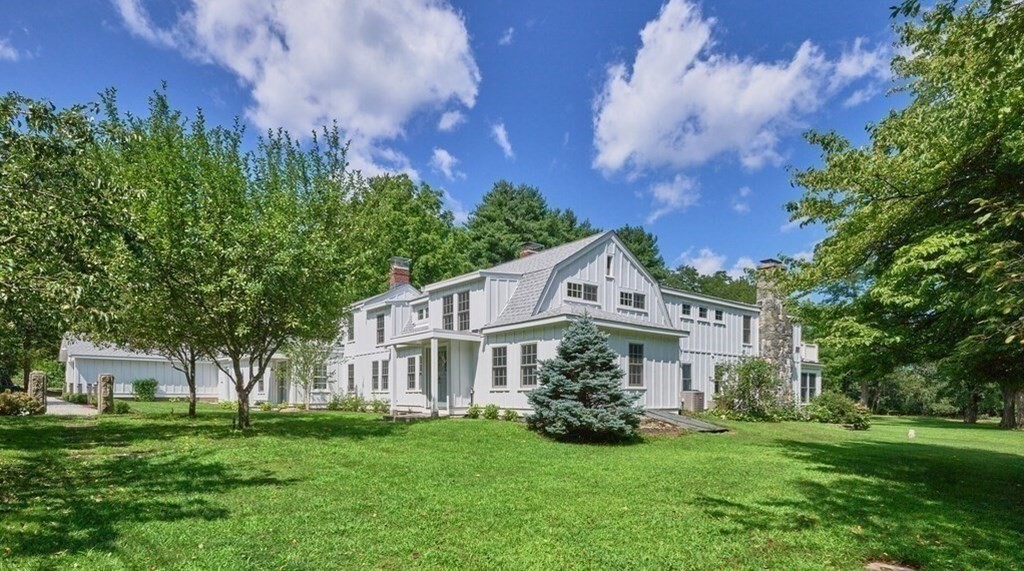 White multi-story home with a stone chimney, broad green lawn, and detached outbuilding.