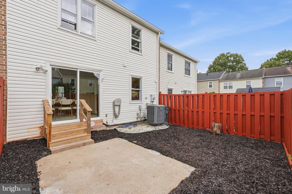Home’s rear patio and fenced yard, with wooden steps, dark mulch beds, and a red privacy fence.