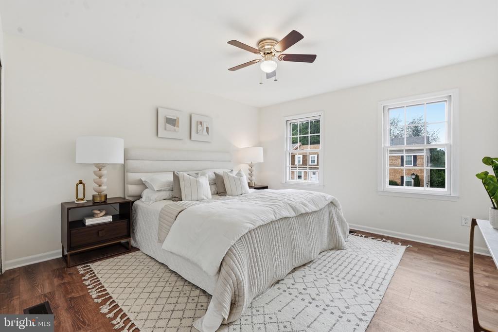 Staged bedroom with a white bed, geometric rug, ceiling fan, and two windows.