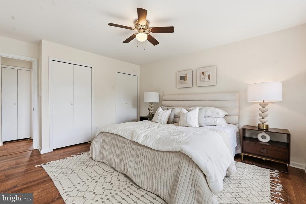 Staged bedroom with a white bed, padded headboard, ceiling fan, and hardwood floors.