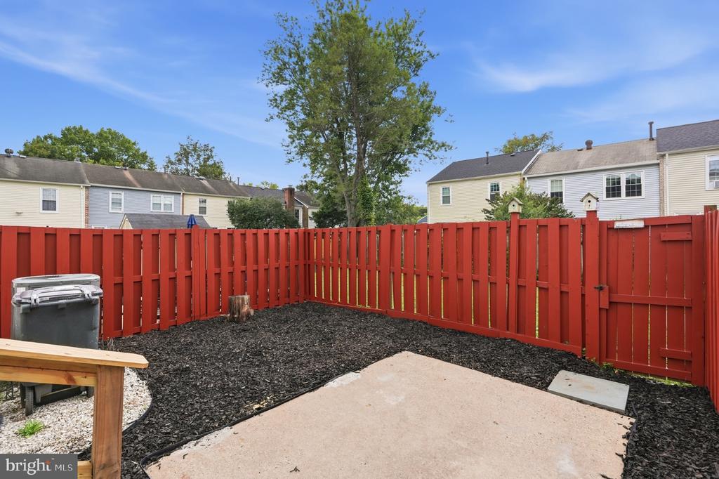Backyard patio bordered by a red privacy fence, with mulch, a grill, and neighboring homes beyond.