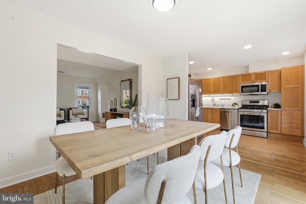 Dining area with a long wood table beside an open kitchen with honey-colored cabinets and stainless-steel appliances.
