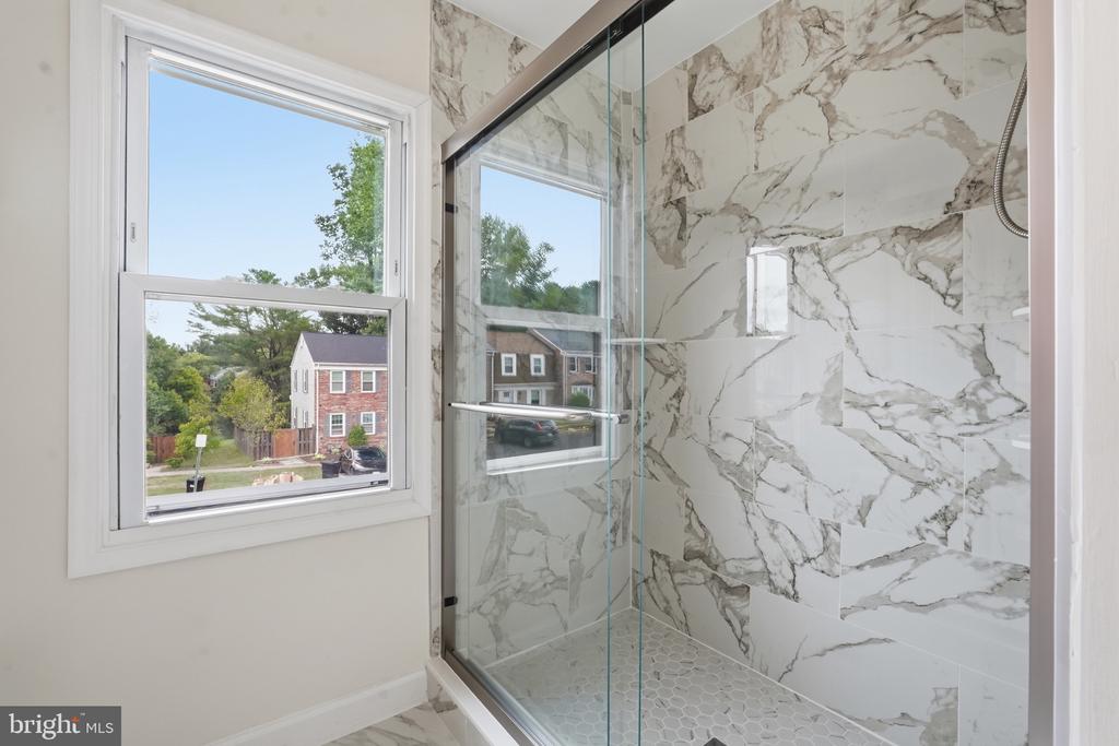 Walk-in shower with marble-look tile, glass enclosure, hexagonal floor, and a window overlooking neighboring homes.
