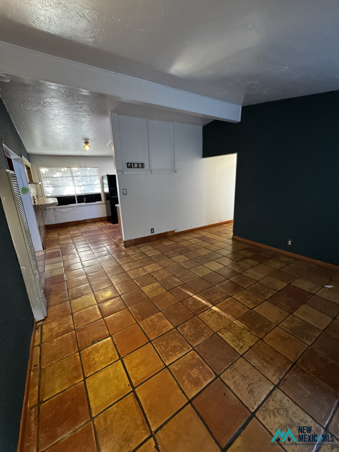 Empty tiled room with a dark teal accent wall and an adjoining kitchen area.
