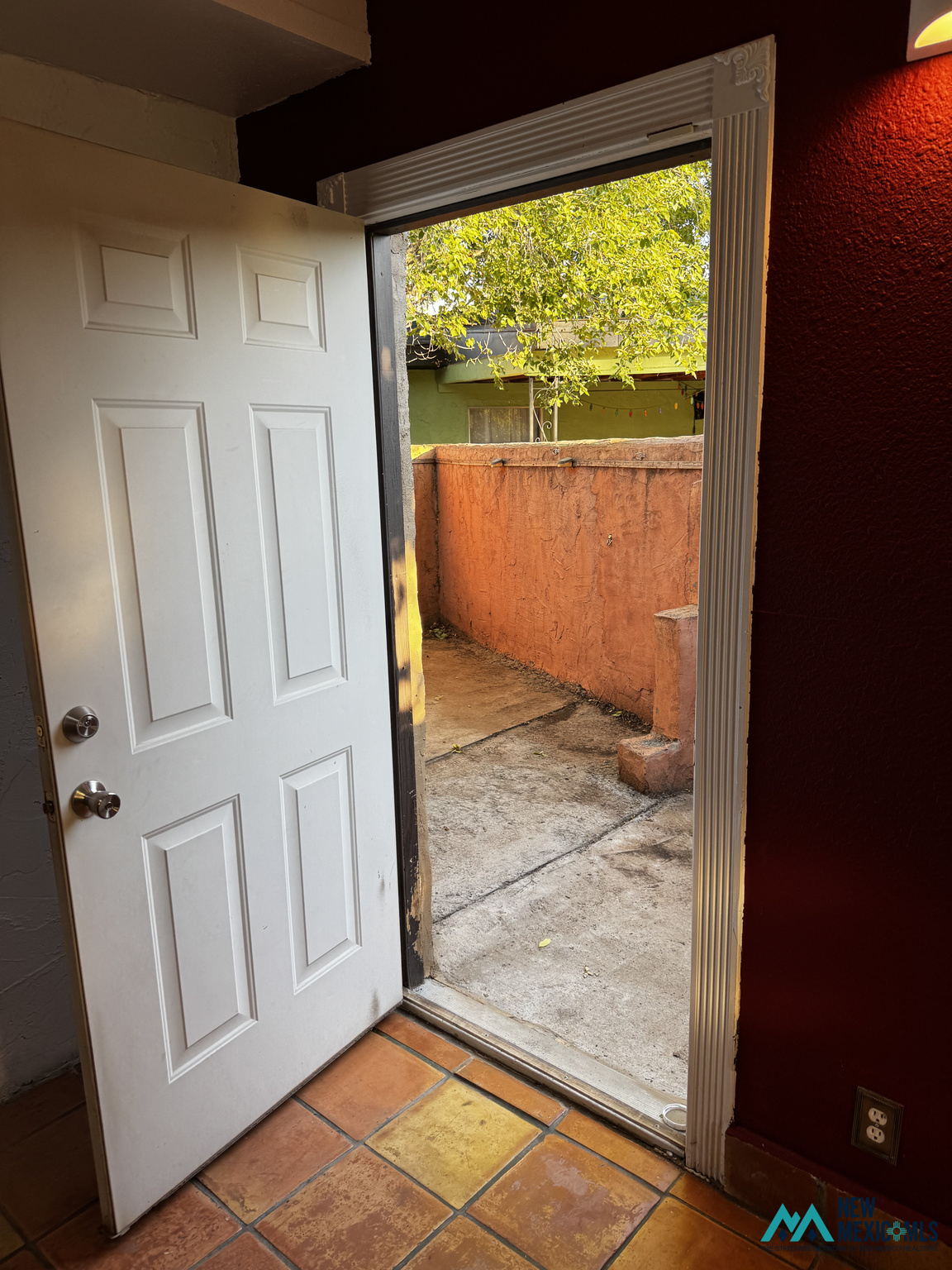 Open doorway to an enclosed patio with an orange stucco wall and leafy branches.