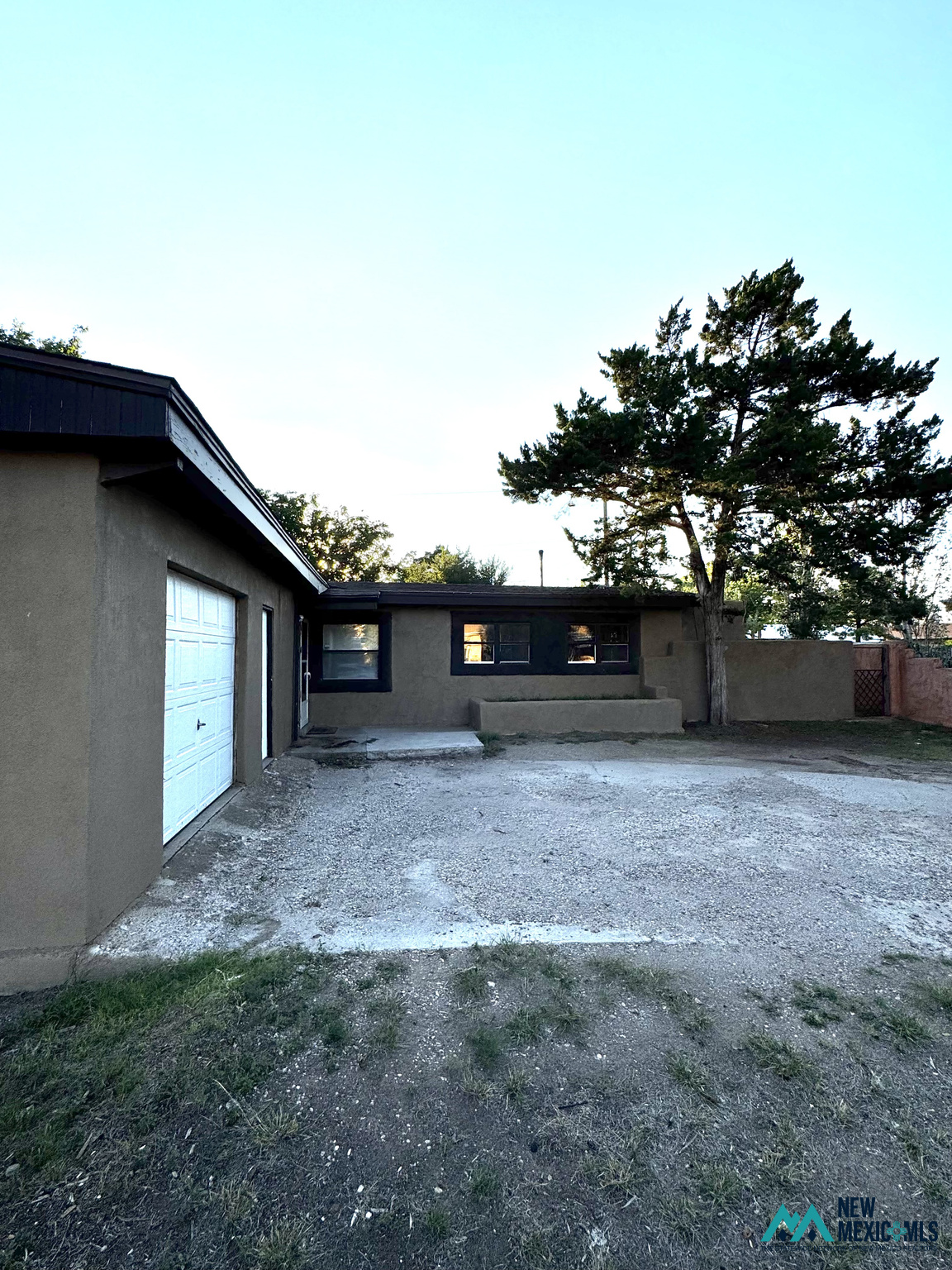 Single-story stucco home with a garage and gravel driveway beneath a large evergreen tree.