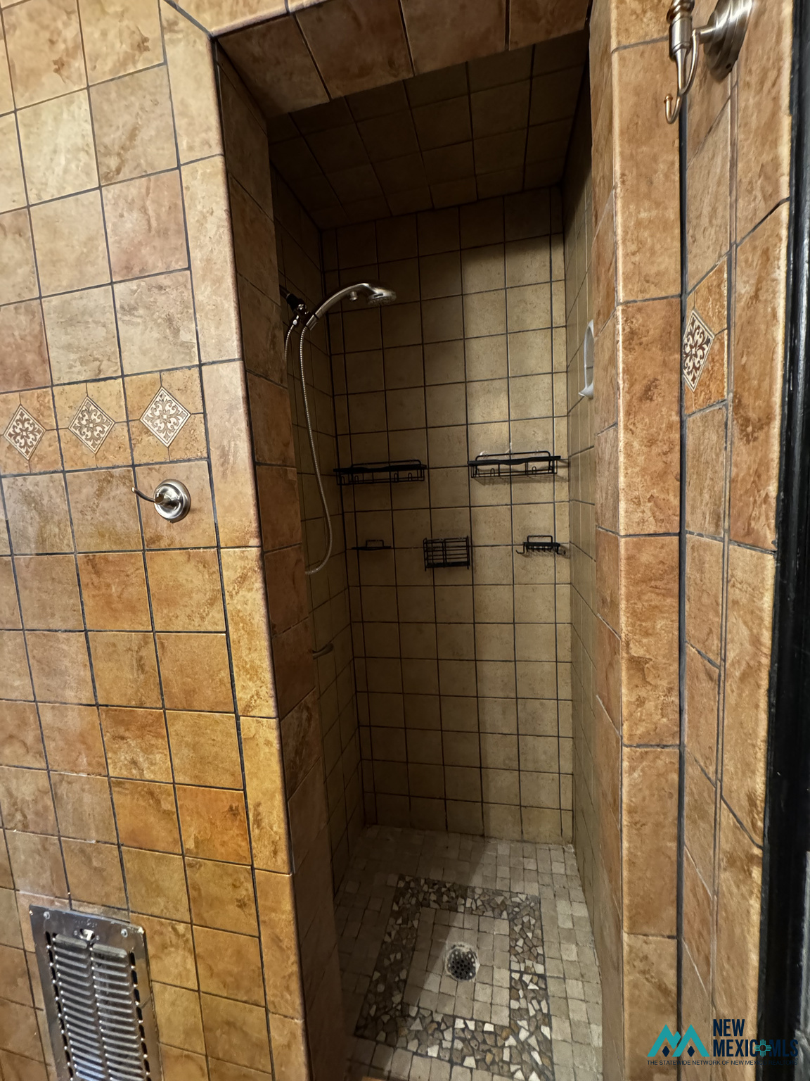Walk-in shower with tan tile, decorative accents, black wire shelves, and a mosaic-bordered drain.