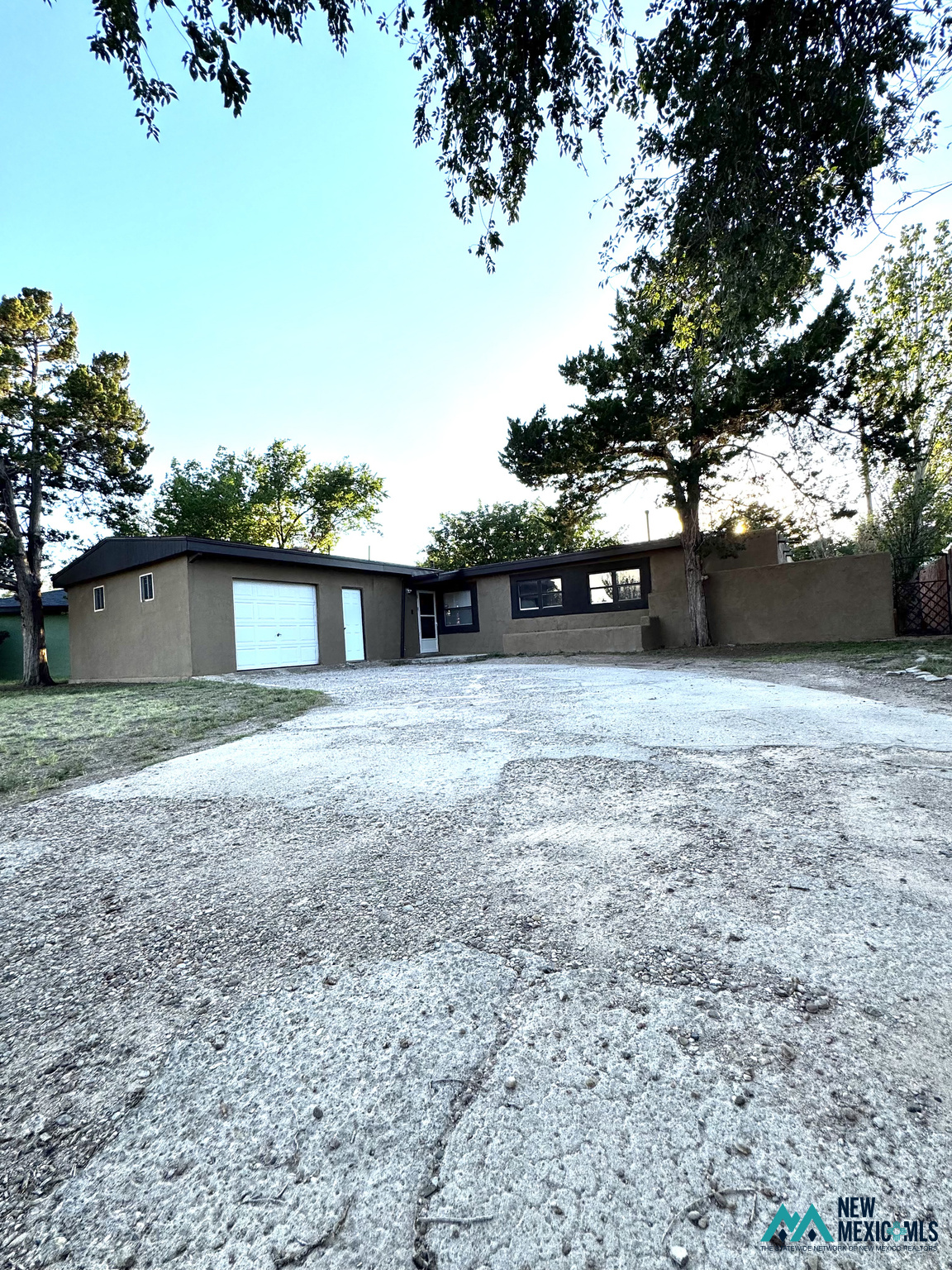 Single-story stucco home with a white garage door, gravel driveway, and mature trees.