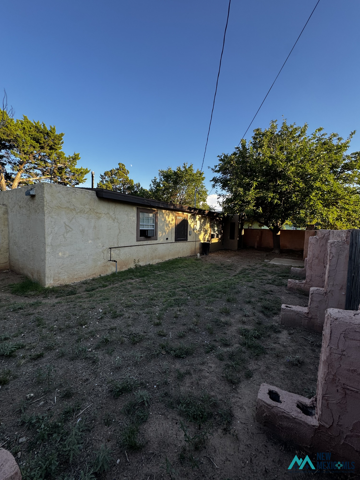 Rear of a single-story stucco house with a dry yard, mature trees, and a low masonry wall.