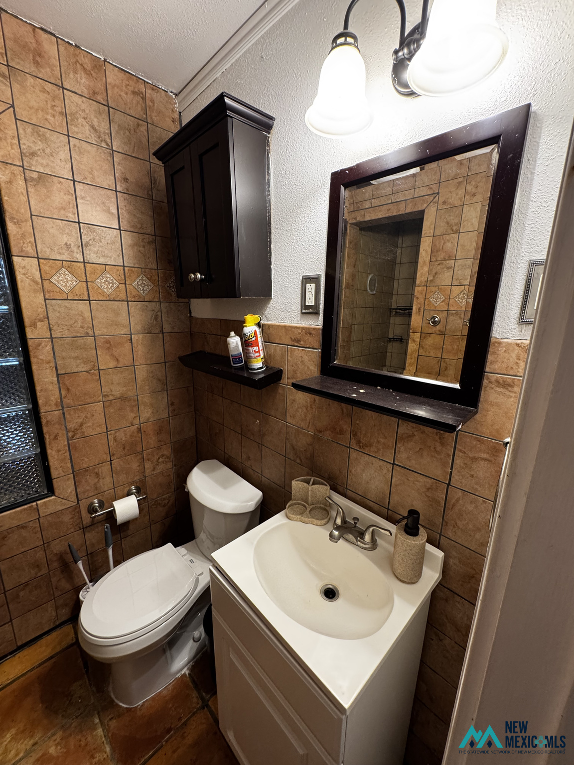 Bathroom with brown tile, a white toilet, and a light vanity beneath a dark-framed mirror.