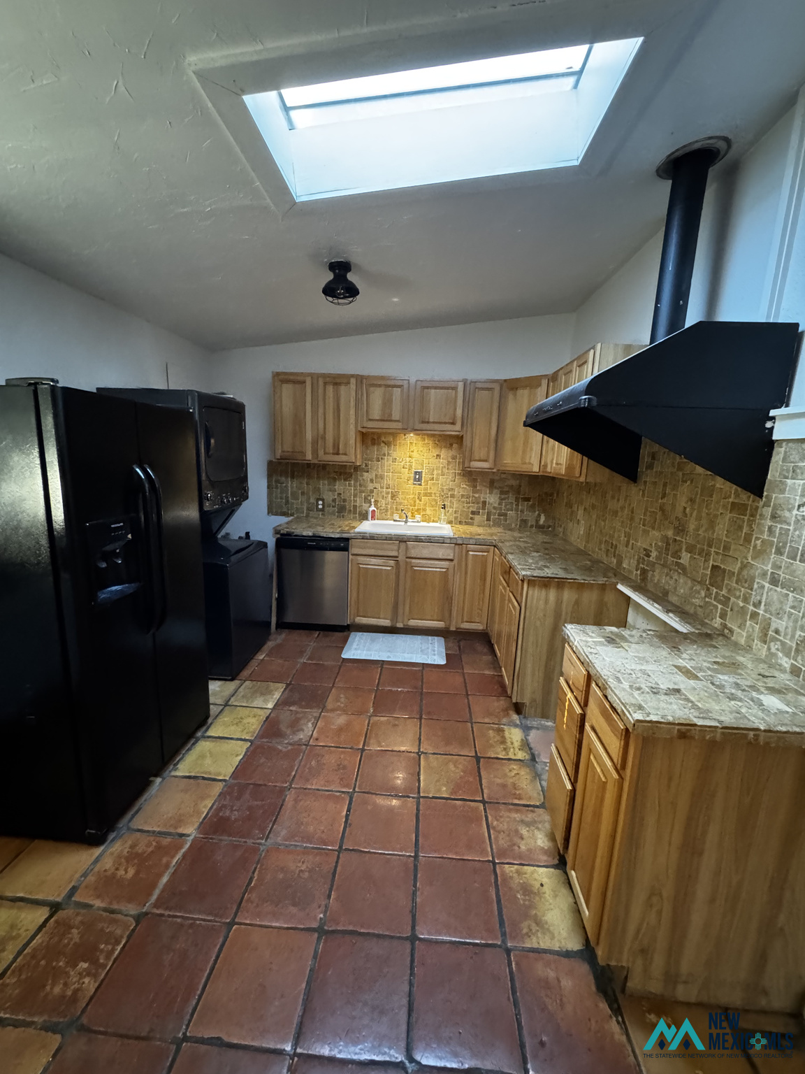 Kitchen with wood cabinetry, black appliances, reddish tile floors, and a large skylight.