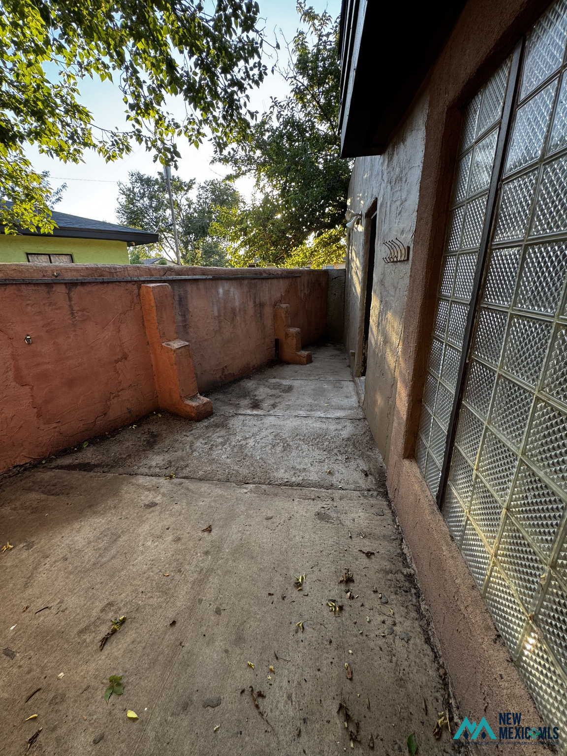 Concrete side patio enclosed by a reddish stucco wall, beside glass block windows and leafy trees.