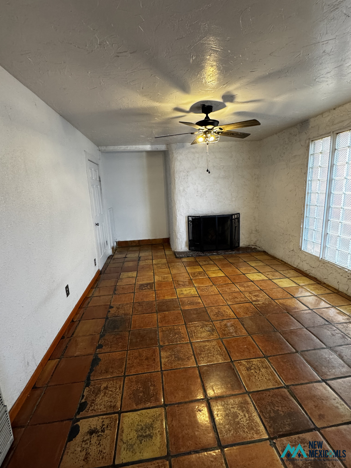 Empty room with terracotta tile floors, a corner fireplace, ceiling fan, and tall gridded windows.
