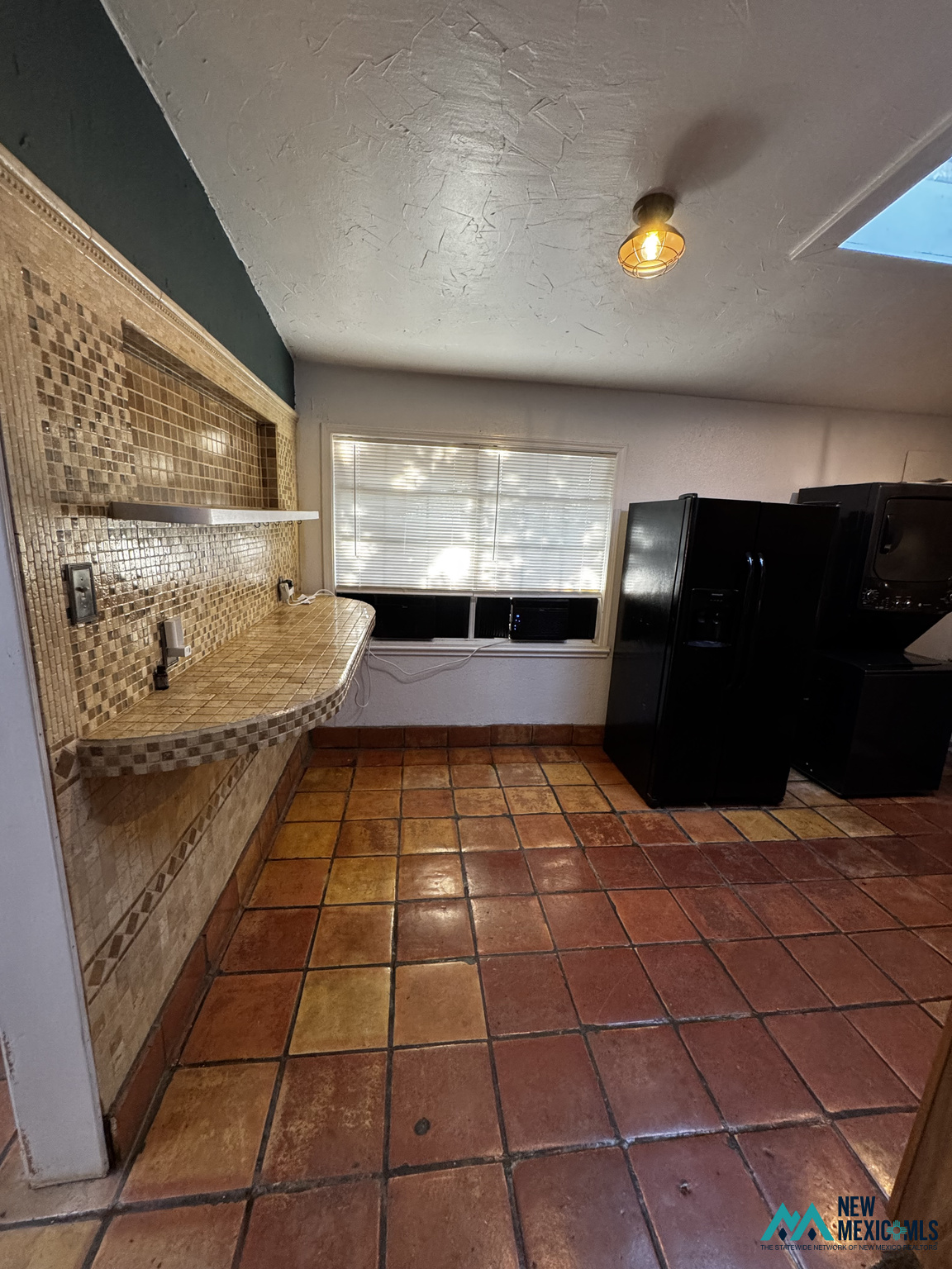 Kitchen with a tiled counter and backsplash, terracotta floor, window with blinds, and black appliances.