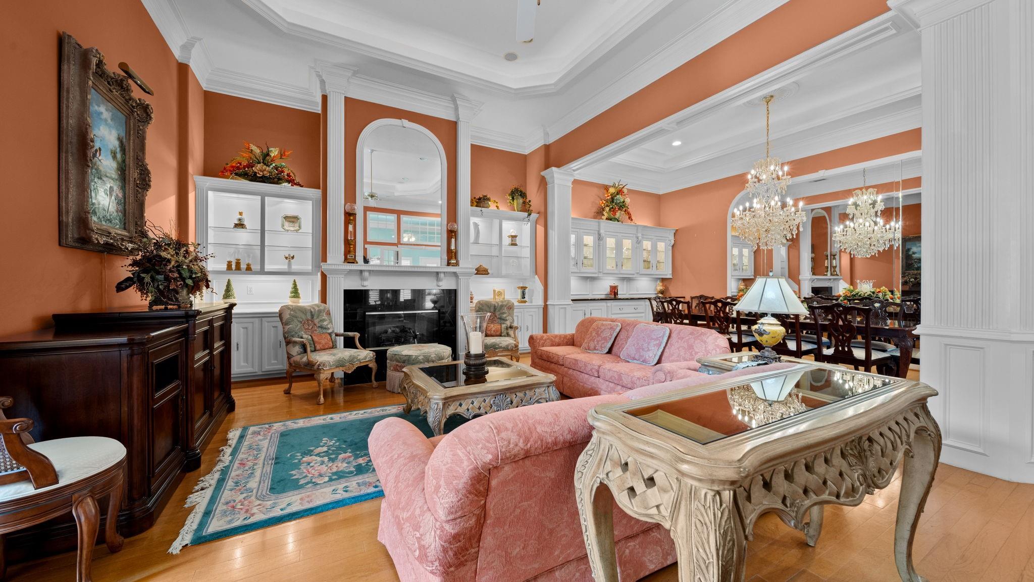 Living room with coral walls, white built-ins, a fireplace, and pink sofas opening to a chandelier-lit dining area.