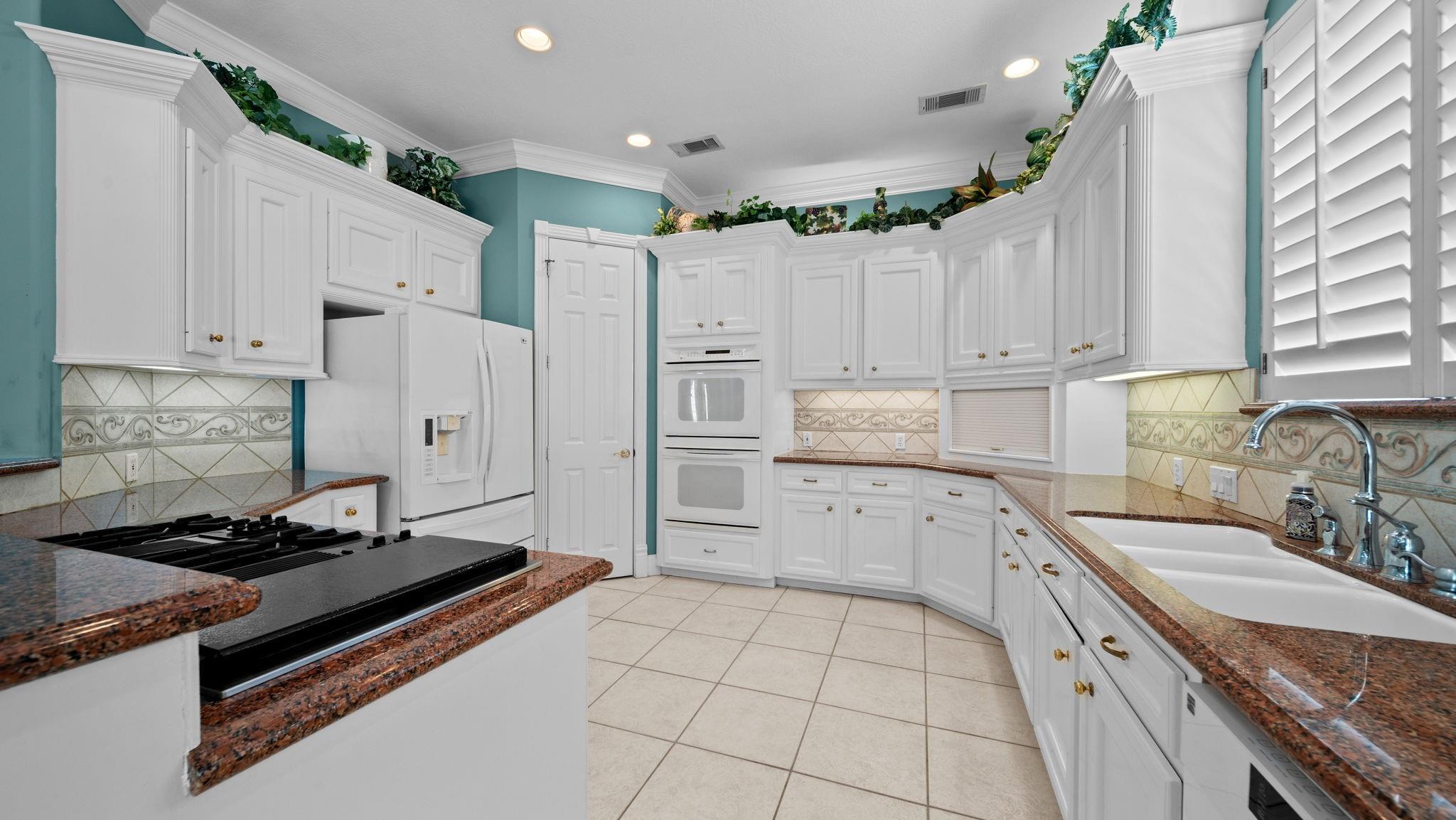 Kitchen with white cabinetry, teal walls, granite counters, double ovens, and a sink beneath shutters.