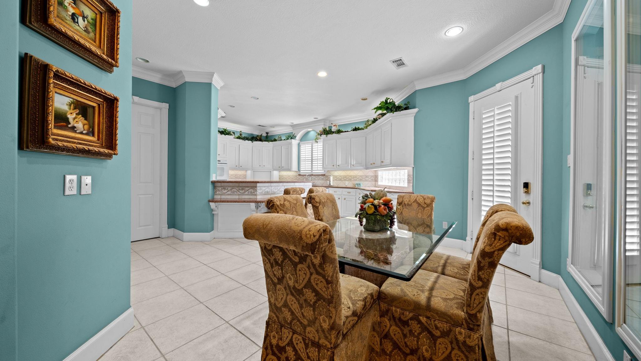 Dining area with patterned chairs and a glass table beside a white kitchen with turquoise walls.