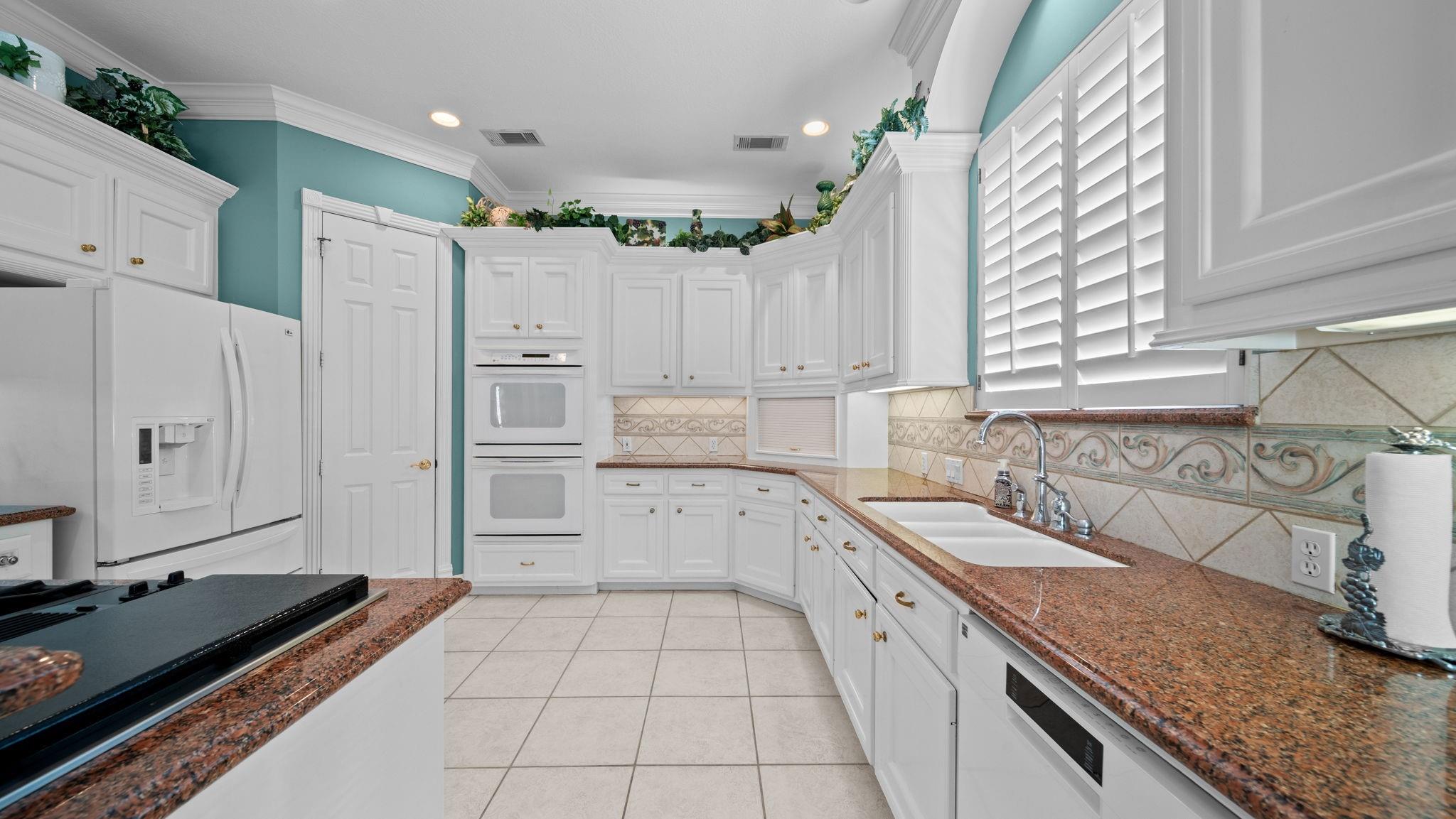 Kitchen with white cabinetry, reddish granite counters, double ovens, and a double-basin sink beneath plantation shutters.