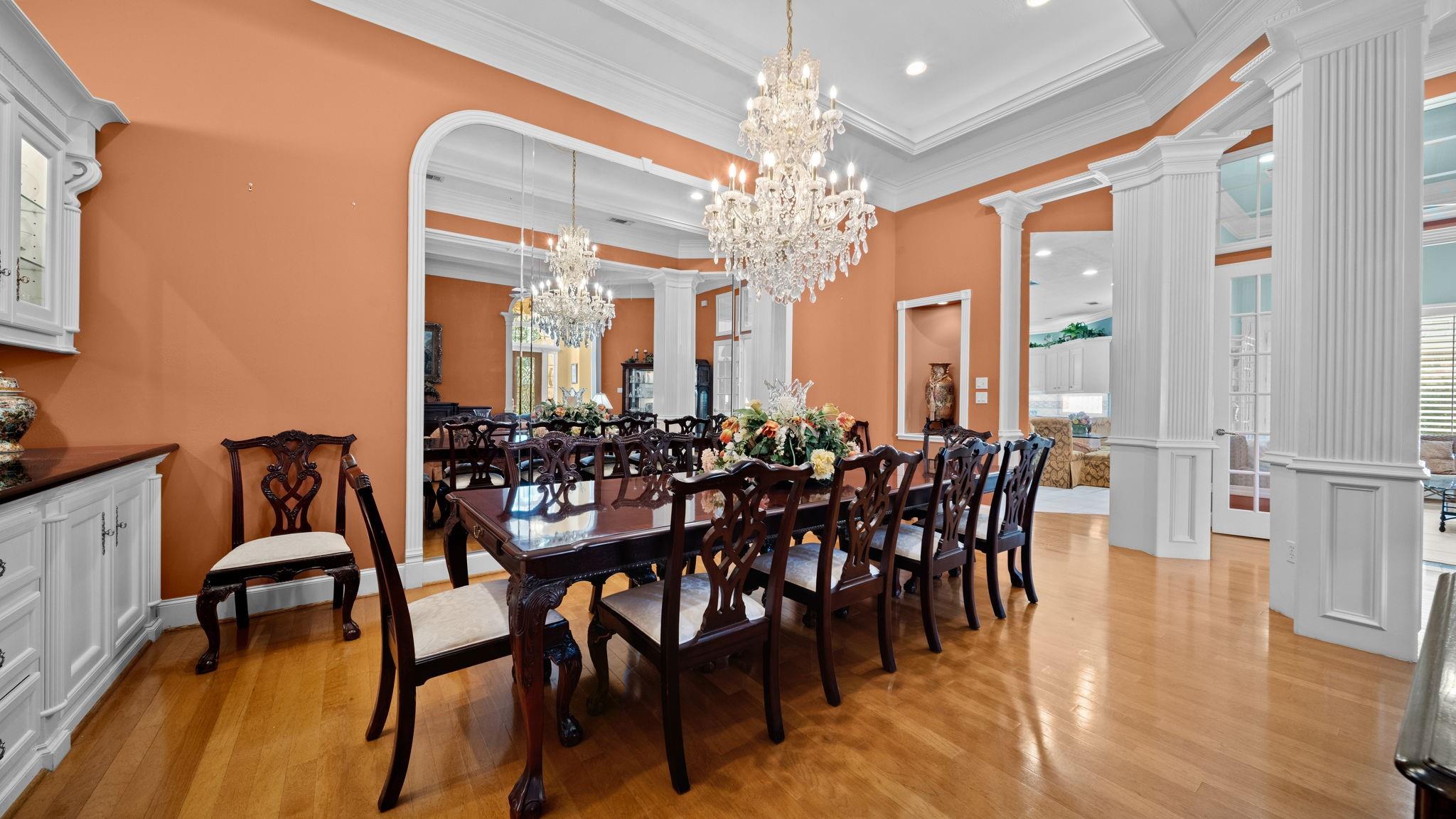 Formal dining room with a long carved table, crystal chandeliers, orange walls, and white columns.