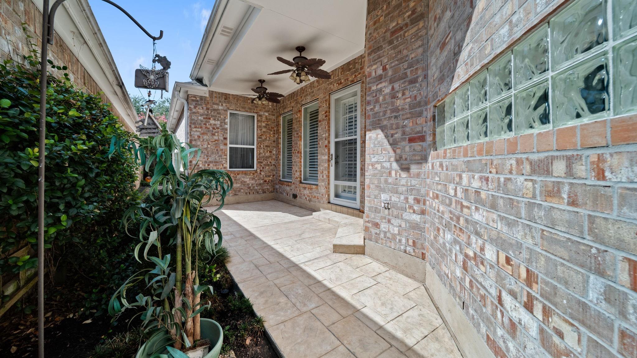 Covered brick patio with tiled flooring, ceiling fans, glass-block window, and garden border.