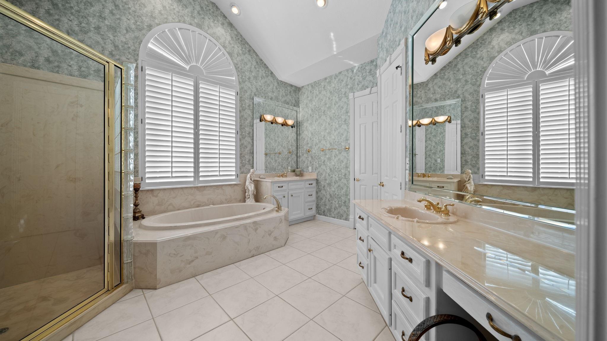 Spacious bathroom with a soaking tub, glass shower, double vanity, and arched shuttered windows.