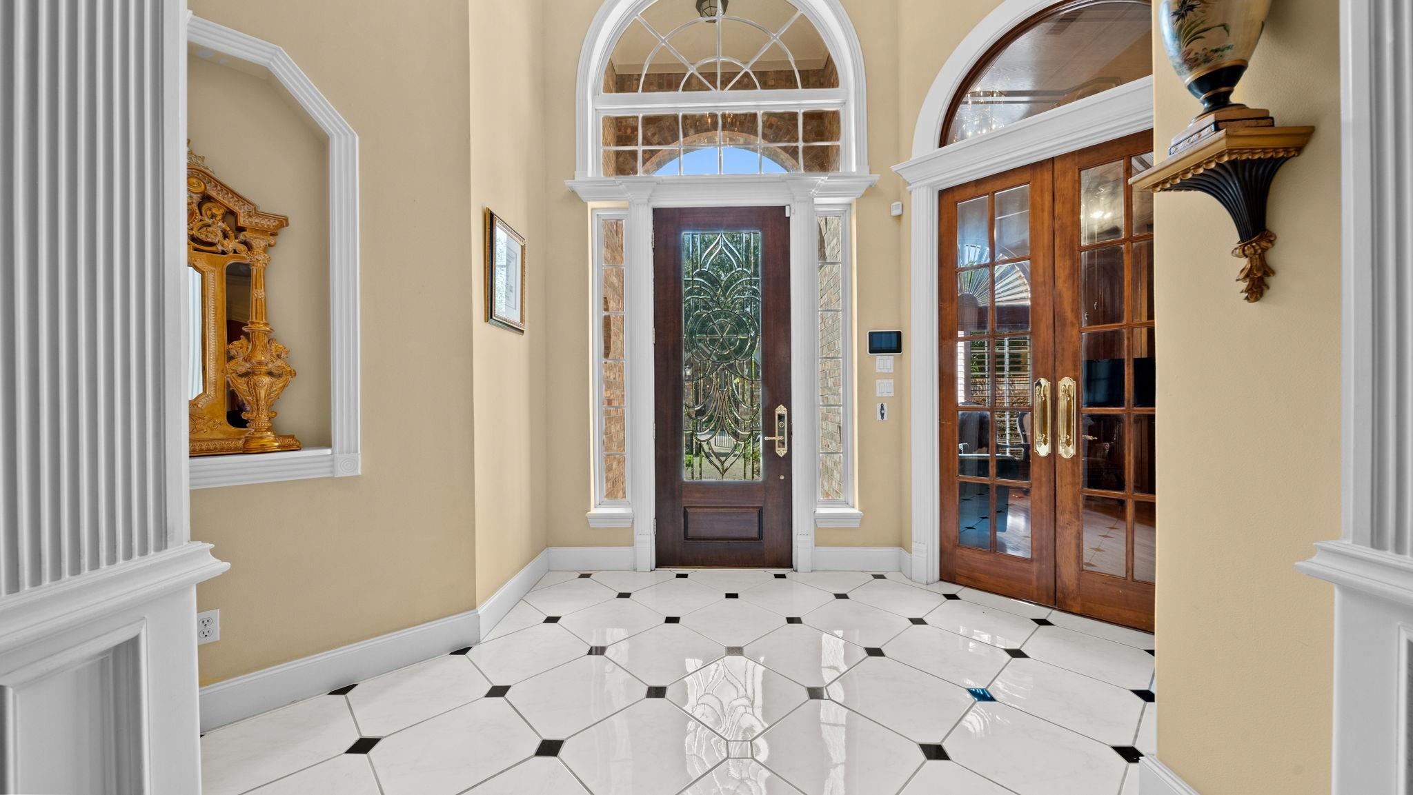 Entry foyer with an ornate glass front door, arched transom, and polished white tile floor with black accents.