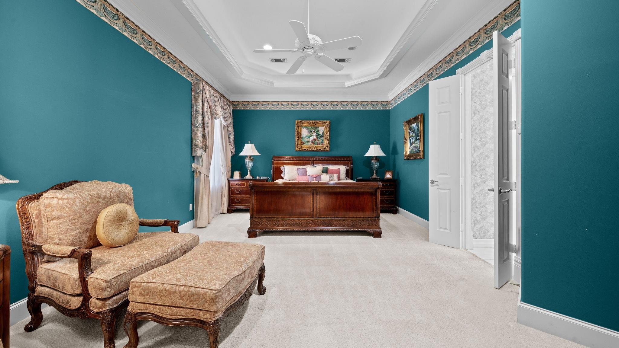 Bedroom with teal walls, dark wood furniture, ornate seating, and a tray ceiling with a ceiling fan.