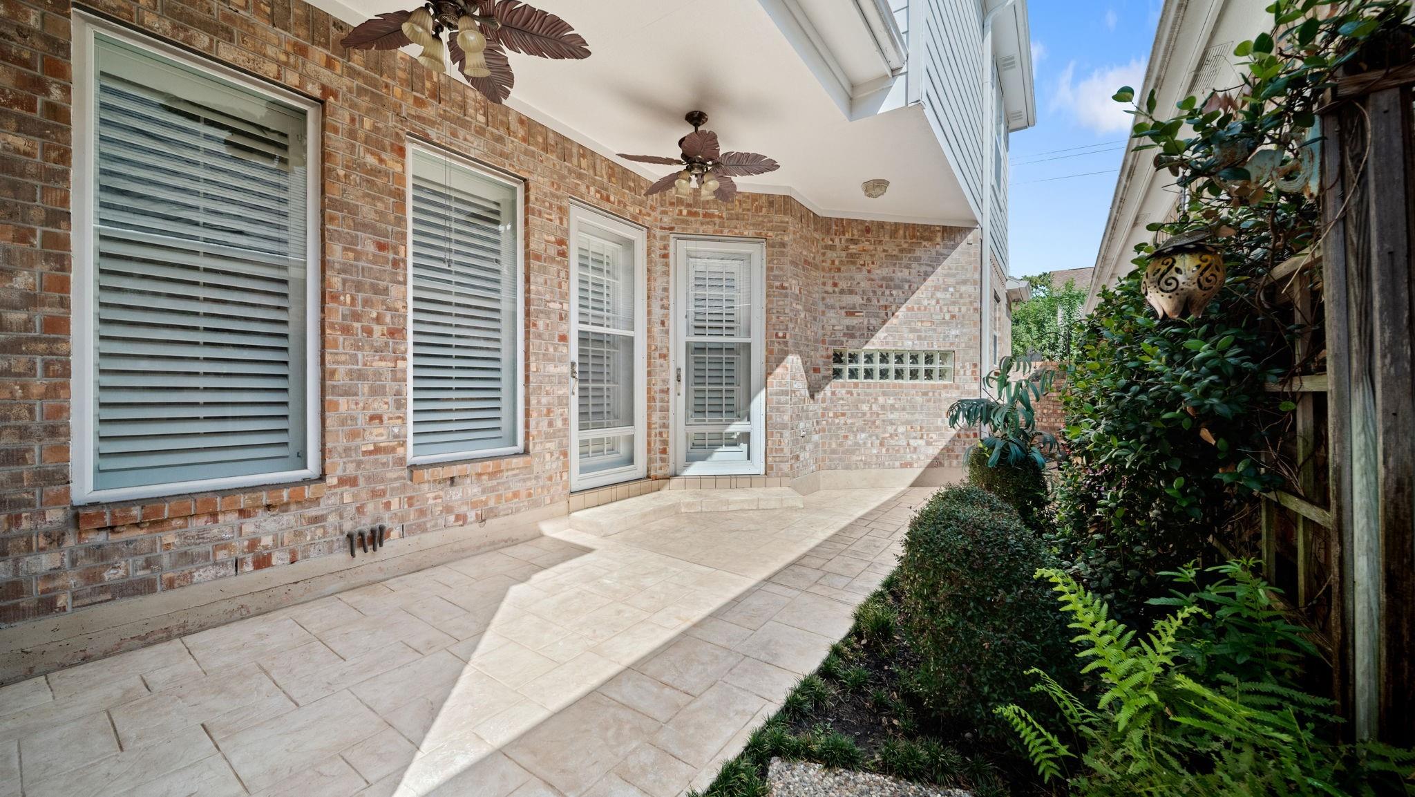 Covered brick patio with stone pavers, ceiling fans, and a leafy garden border.