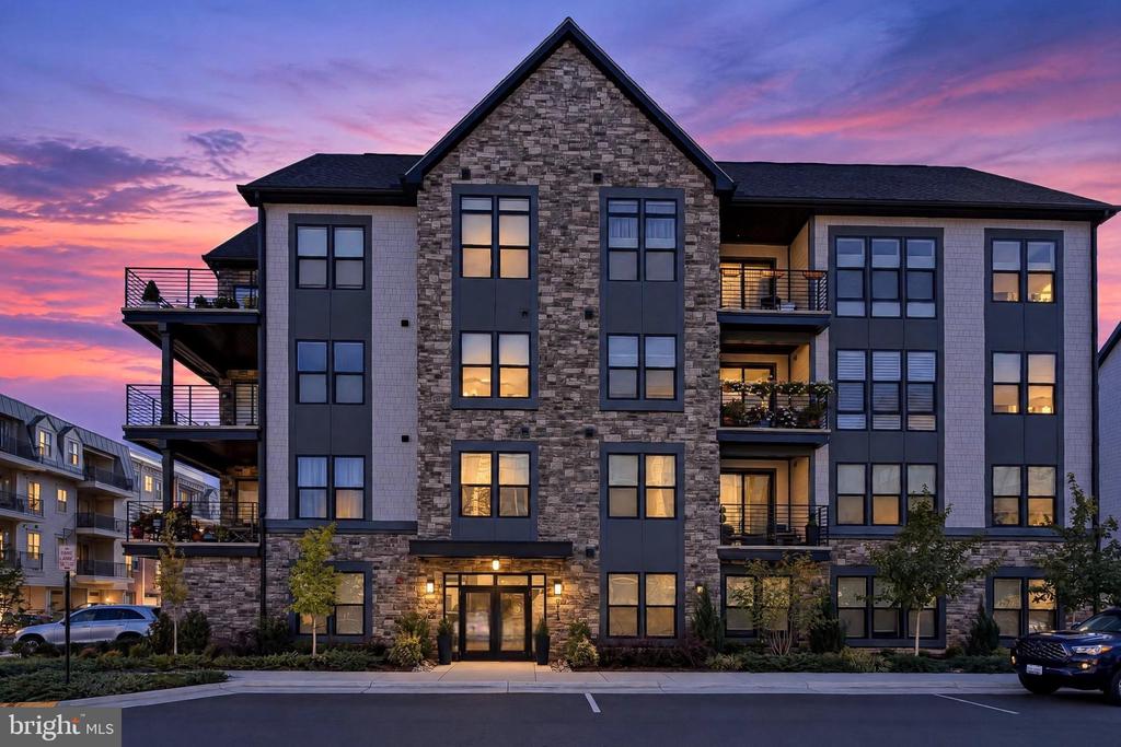 Modern multi-story residential building with stone facade, balconies, and illuminated windows at dusk.