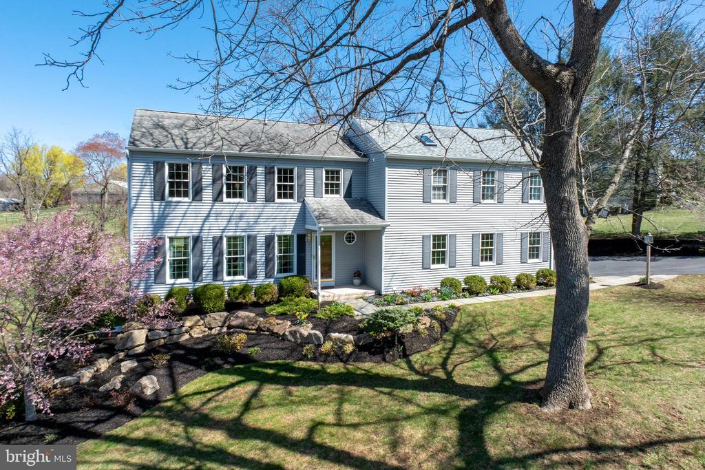 Two-story gray house with dark shutters, a stone garden wall, and a large tree in the foreground.