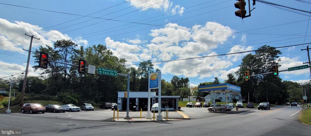Commercial gas station at a tree-lined intersection, with a blue-and-yellow canopy and parked cars.