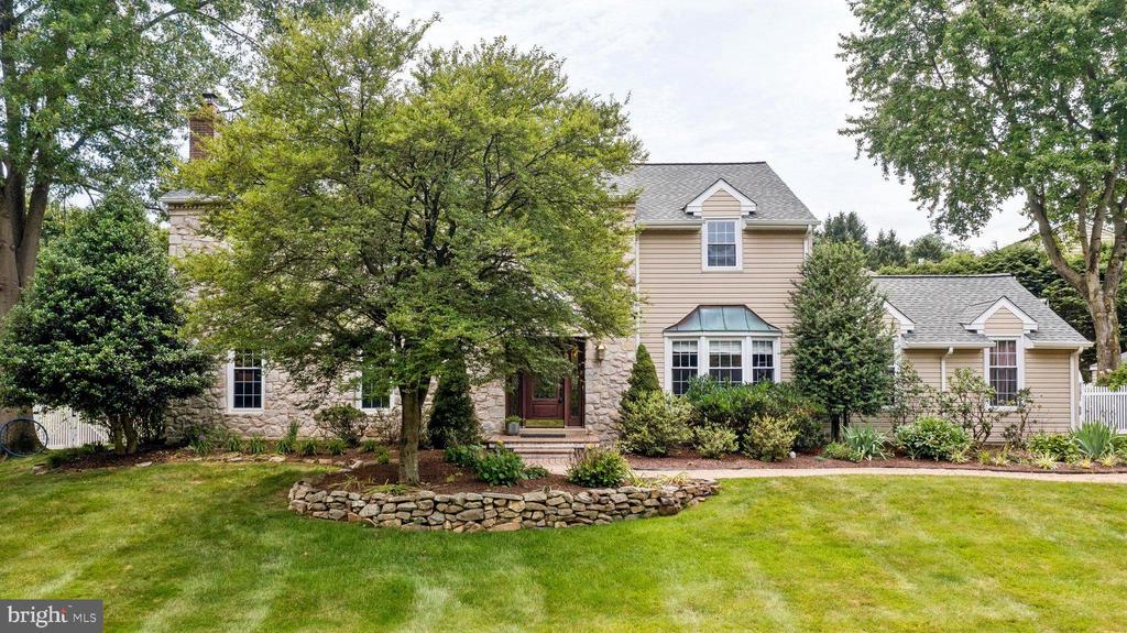 Two-story beige-and-stone home framed by mature trees, with landscaped gardens and a broad lawn.