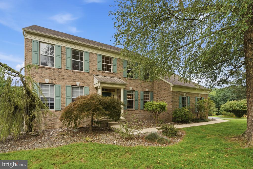 Two-story brick house with pale green shutters, landscaped shrubs, and a lawn beneath mature trees.