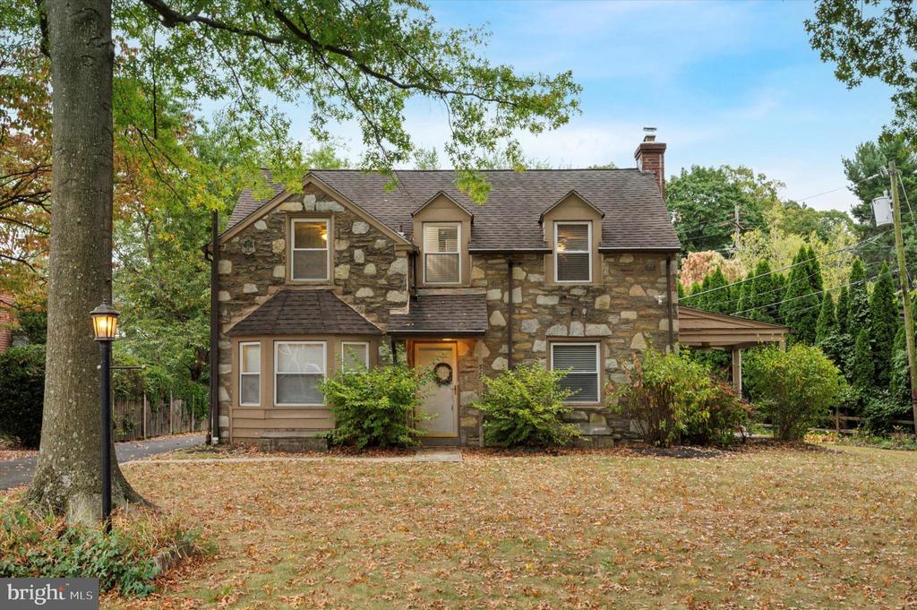 Stone house with dormer windows, a covered carport, and a leaf-strewn front lawn.
